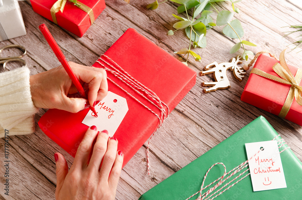 Φωτογραφία Woman's hand writing a Merry Christmas tag with a red marker for a Christmas gift wrapped with red paper on a wooden table