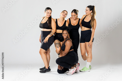 Full photograph of a group of 5 girls of different ethnicities wearing black sportswear posing against a white background. The black girl is seated.Concept of women in the sports world.