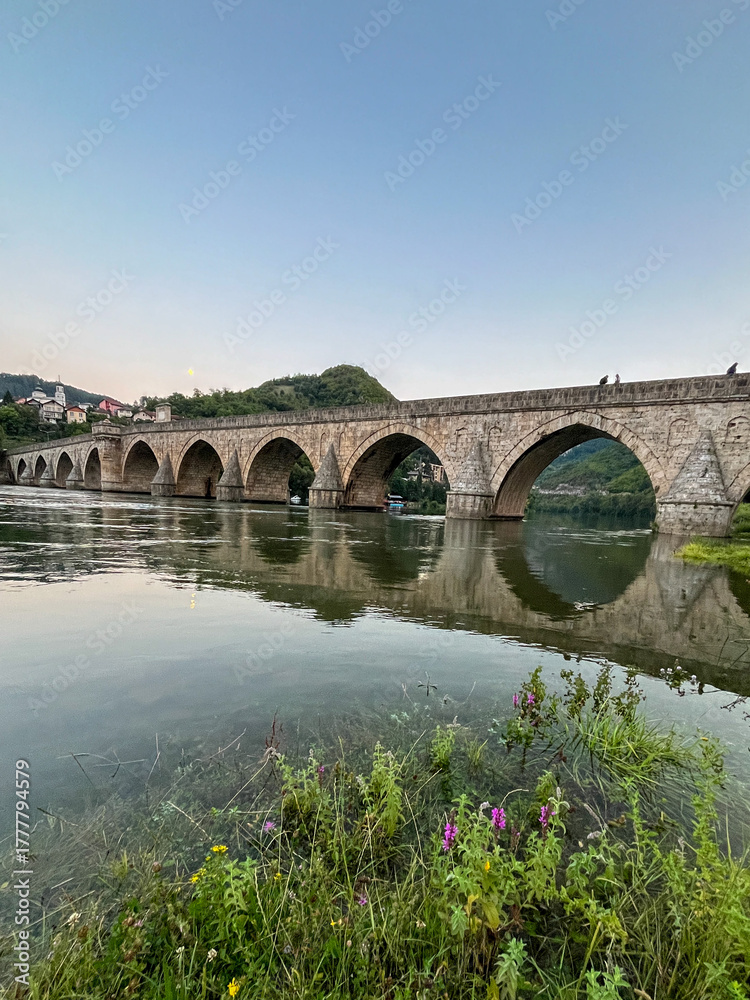 Fototapeta premium Historic stone bridge with illuminated arches over the Drina River at dusk.