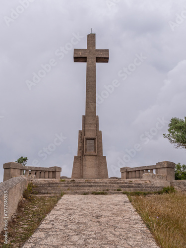 Puig des Mila, Sant Salvador, Mallroca, large stone cross monument stands at the end of a stone pathway, surrounded by grass and trees under a cloudy sky, Majorca