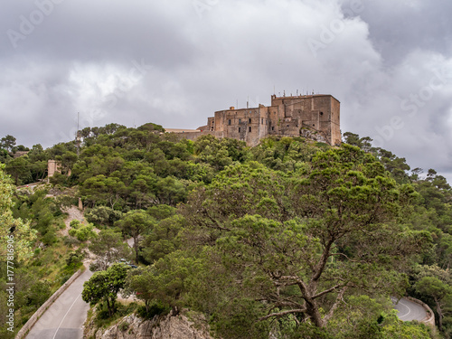 Santuari de Sant Salvador, Mallorca, historic castle perches atop a lush hill, surrounded by dense green trees, under a cloudy sky, Majorca