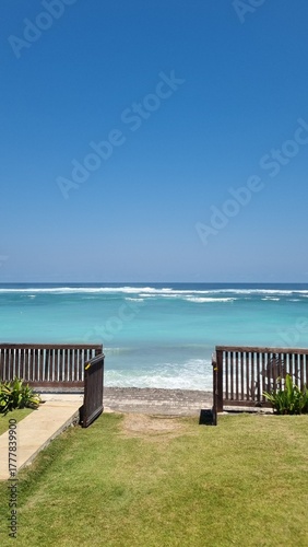 Beautiful tropical beach view with turquoise ocean, clear blue sky, wooden fence and open gate leading to the sea. Peaceful summer day at seaside, perfect vacation and travel destination.