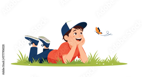 Happy young boy in a cap lying on green grass, looking up with wonder at a beautiful orange butterfly flying in the sky.