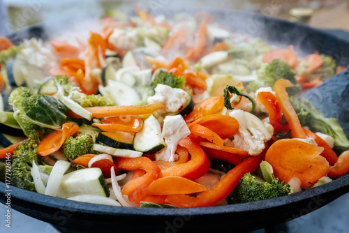 A vibrant mix of steaming stir-fried vegetables including carrots, broccoli, zucchini, cauliflower, and bell peppers, cooking in a large black pan outdoors.