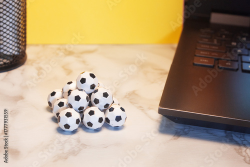 Pyramid of small soccer balls next to a laptop on a marble surface with a bright yellow background