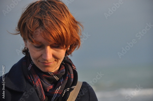 Young woman with short red hair smiles softly while standing on the beach during a cloudy day