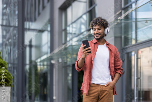 Young Indian man wearing headphones walking on the street and using his mobile phone