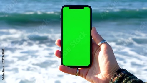 A person holds a smartphone with a green screen display against the backdrop of a beautiful blue ocean with gentle waves crashing on the shore perfect for showcasing mobile apps or travel content.