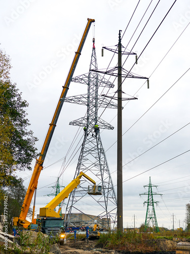 Workers installing high-voltage power line tower using cranes and lifts at construction site