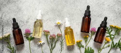 Border of various glass dropper bottles with essential oil or serum on rustic background with wild flowers, natural cosmetic products, top view, flat lay