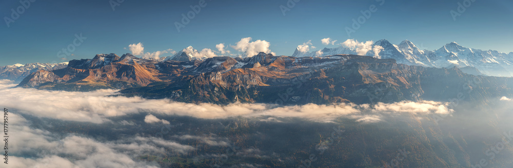 Fototapeta premium Panoramic aerial view of the Swiss Alps with snow-capped peaks and low clouds at sunset. Bright evening light and clear blue sky over rugged alpine mountains in Switzerland. Top view. Landscape