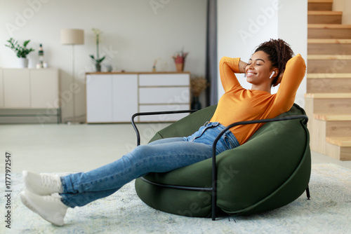 A young woman relaxes in a modern chair at home, wearing earbuds and smiling. She is dressed casually in an orange sweater and jeans, enjoying a moment of peace.