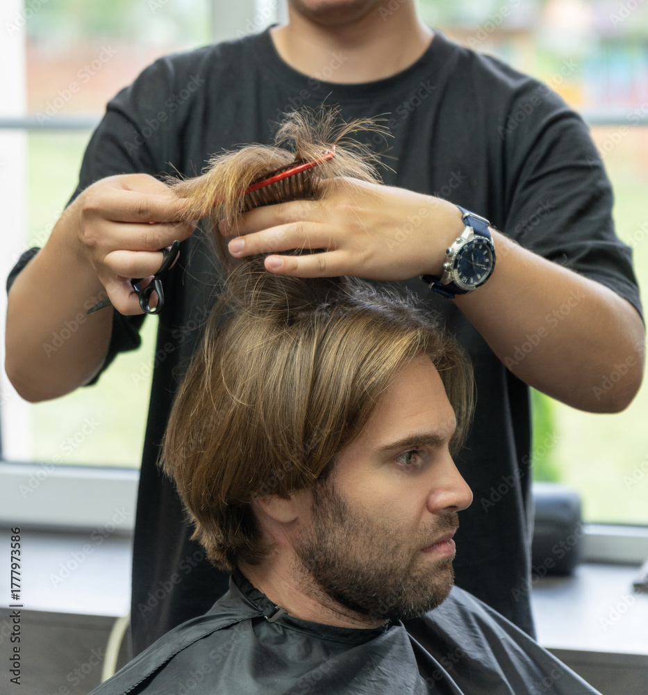 Fototapeta premium A hairdresser uses a pair of scissors to cut a male client's long blonde hair.