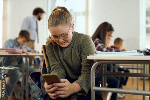 Smiling elementary student using mobile phone while having a class with her friends at elementary school