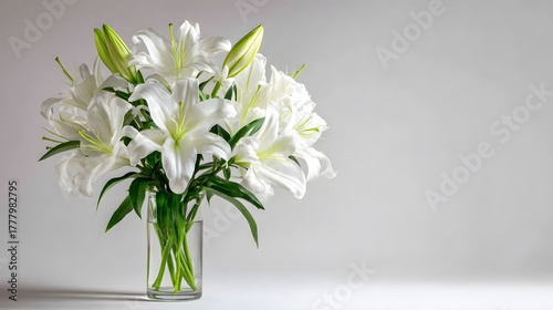 Abundant bouquet of pure white lilies stands elegantly in a clear glass vase against a neutral background