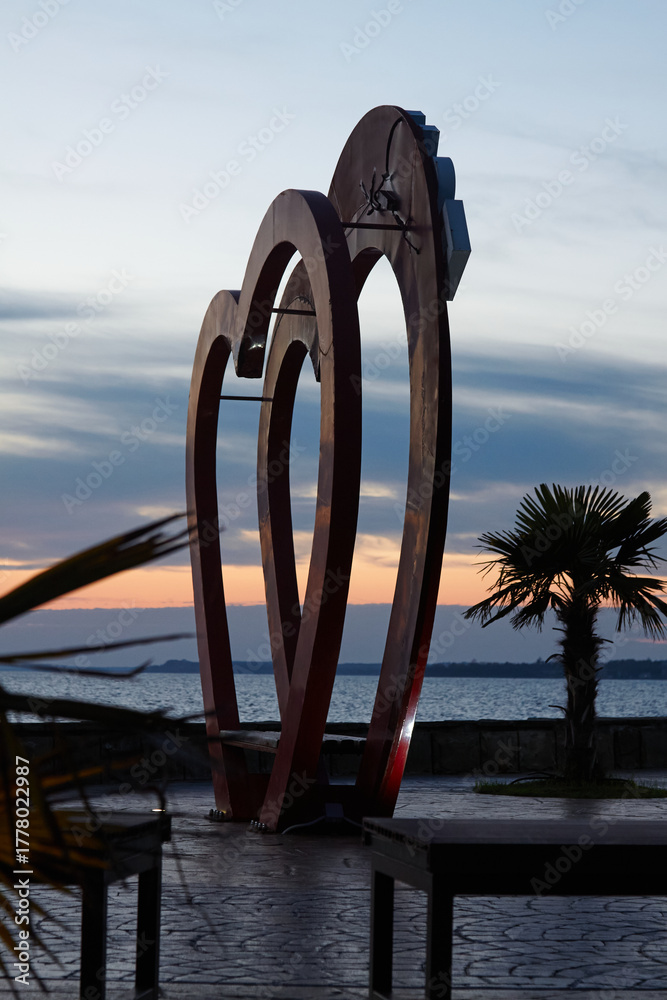 Fototapeta premium Large heart-shaped sculpture stands prominently against a colorful sunset sky, surrounded by palm trees and a serene ocean view, creating a romantic and picturesque atmosphere for visitors