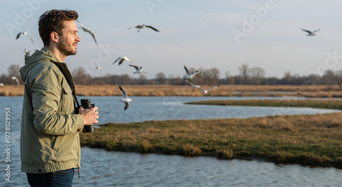 thoughtful man birdwatching with binoculars near river under bright sky in tranquil landscape