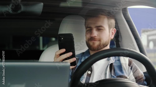 Smiling young man sitting in the car and looking at phone screen. Slow motion
