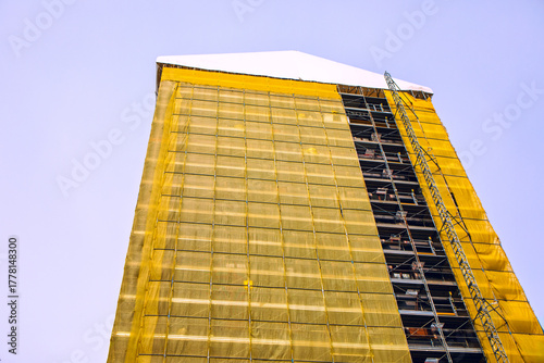 Towering building under construction stands tall, draped in yellow scaffolding netting against blue sky. Its peaked white roof contrasts sharply with the exposed metal frameworks and ladders below