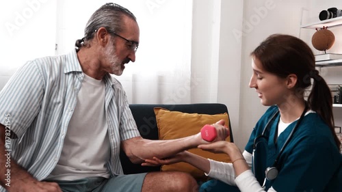 A young female nurse cares for an elderly man sitting while his grandfather lifts dumbbells to build muscle and stretch in a nursing home.
