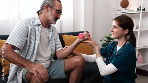 A young female nurse cares for an elderly man sitting while his grandfather lifts dumbbells to build muscle and stretch in a nursing home.
