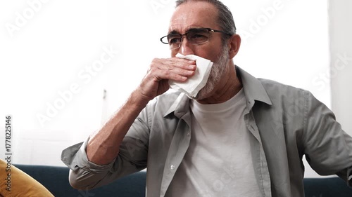 Elderly man sitting on sofa sick using tissue paper sneezing coughing feeling unwell having cold at home. Health concept.
