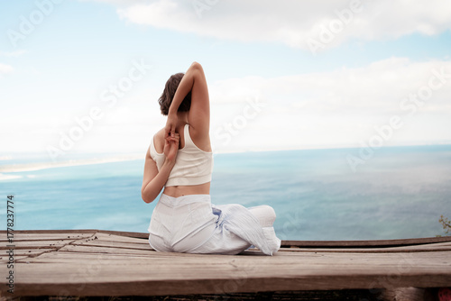Back view of woman in sportswear sitting in Gomukhasana Cow Face Pose, stretching shoulders and arms. She is enjoying serene ocean view and peaceful nature during yoga practice