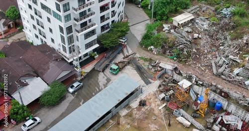 Seen from above, long steel beams are being unloaded from a flatbed by workers using a crane truck in a yard located right beside a modern apartment building.