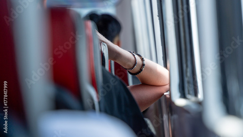 Fotografía A close-up of a passenger's arm, wearing a beaded bracelet, resting on the back of a red bus seat while they look thoughtfully out the window during travel
