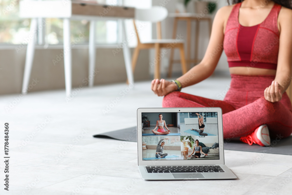 Fototapeta premium Sporty young African-American female coach with yoga mat and laptop meditating at home. Concept of online training