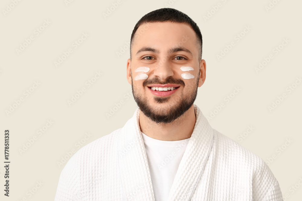 Obraz premium Young man in bathrobe with cream smears on cheeks against white background