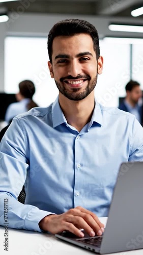A man in a blue shirt gestures towards the viewer, working on a laptop in an office setting
