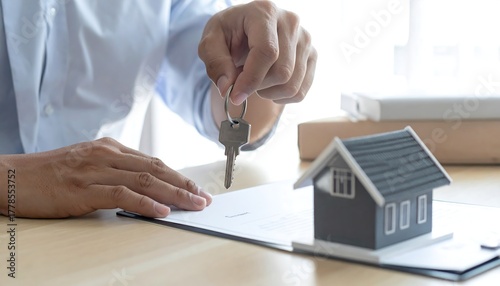 A person holding keys over a clipboard next to a small gray house model on a light wood table