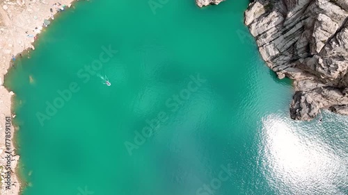 Direct aerial view of the brilliant turquoise water of Kel-Suu Lake showing a small boat, sun glare, and a rocky shoreline with a few people and tents