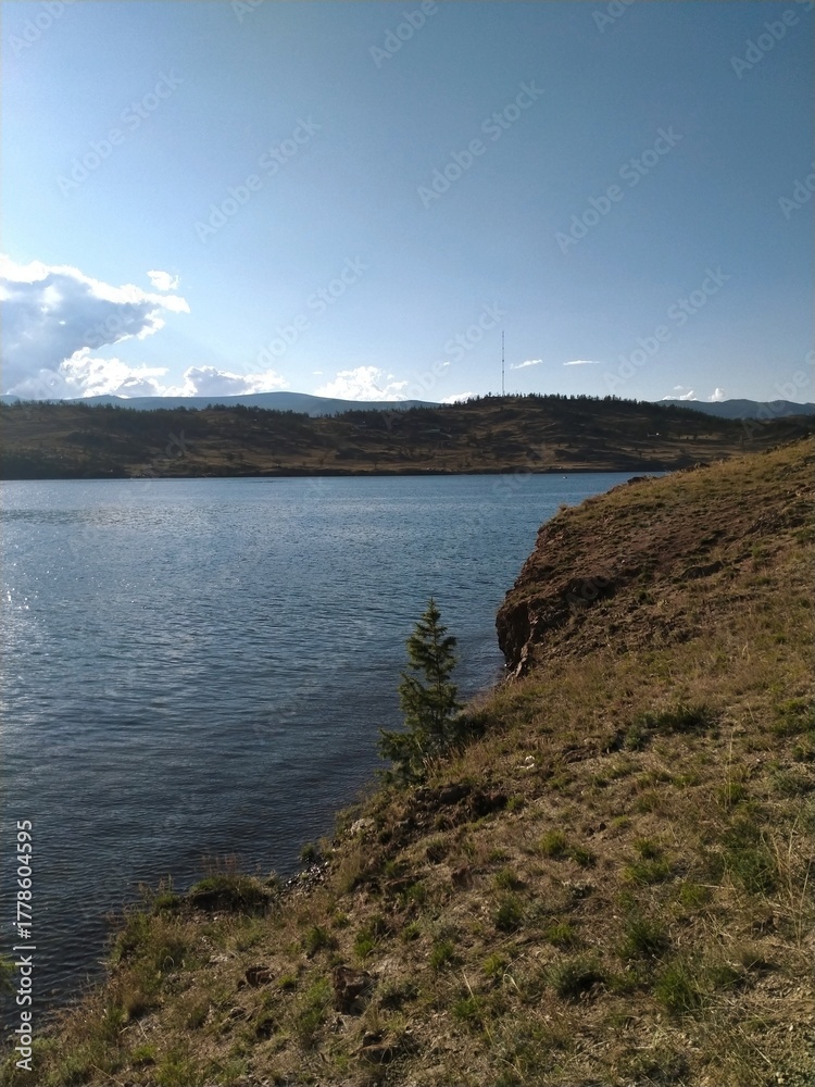Fototapeta premium A sunny slope over calm water and distant hills. Clear sky, natural scenery, summer atmosphere and tranquility.