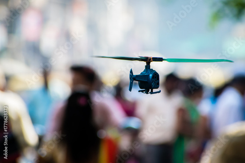 Fototapeta Naklejka Na Ścianę i Meble -  A small, remote-controlled toy helicopter flies in the air, sharply focused against a soft, blurred background of people and a busy street crowd.