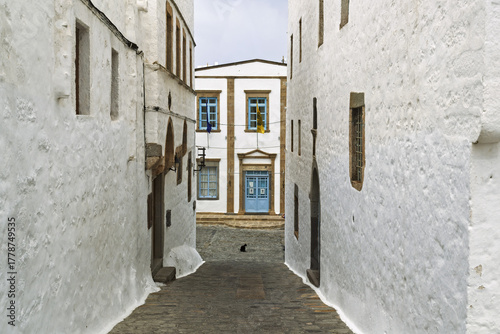 Fototapeta Naklejka Na Ścianę i Meble -  Narrow cobblestone alleyway between tall whitewashed buildings with small windows in the Monastery of Saint John the Theologian on the island of Patmos, Greece.