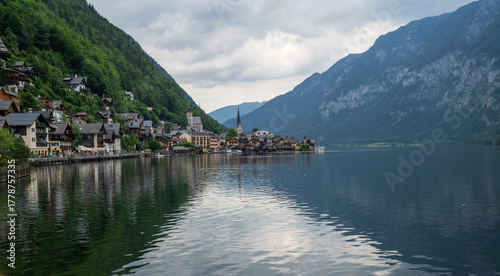 Lakeside village of Hallstatt in the Austrian Alps with traditional alpine houses, mountain slopes covered in dense forest, and church towers reflected in the calm waters of lake under cloudy skies.