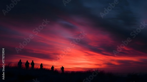Fototapeta Naklejka Na Ścianę i Meble -  A group of silhouetted figures stands on a hill observing a dramatic and vibrant sunset
