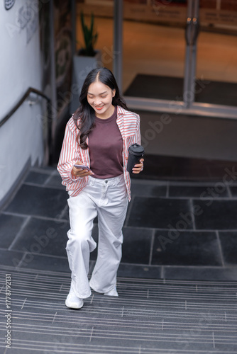 Fotografie Young woman walking upstairs checking smartphone with coffee