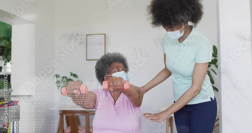 Senior woman wearing mask holding pink dumbbells as caregiver wearing polo guiding movement at home