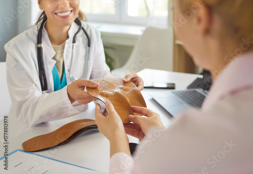 Bild auf Leinwand Smiling doctor showing a pair of orthopedic insoles to woman patient during medical consultation in clinic