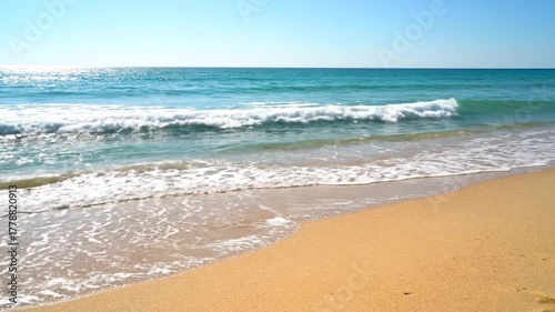 Coastal view features waves breaking on a sandy shore under a bright, clear blue sky