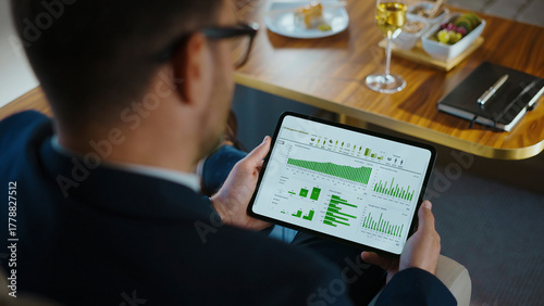 Businessman reviewing a data management dashboard with green charts and graphs on a digital tablet, holding the device while sitting at a table with a drink and notepad