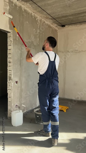 A bearded man in a work uniform is priming the walls after applying gypsum plaster. 