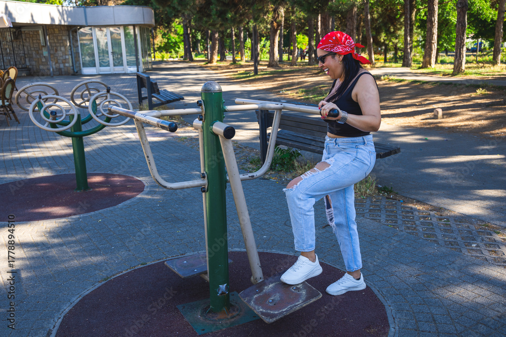 Fototapeta premium A woman exercises on outdoor fitness equipment in a sunny park surrounded by trees. The setting shows an urban recreational area with benches and fitness machines under natural daylight.