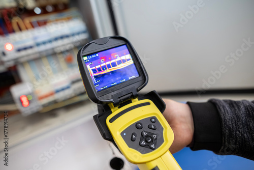 An electrician's hand holds a thermal imaging camera to inspect an open electrical panel, checking for overheating components, hotspots, and potential faults during maintenance.