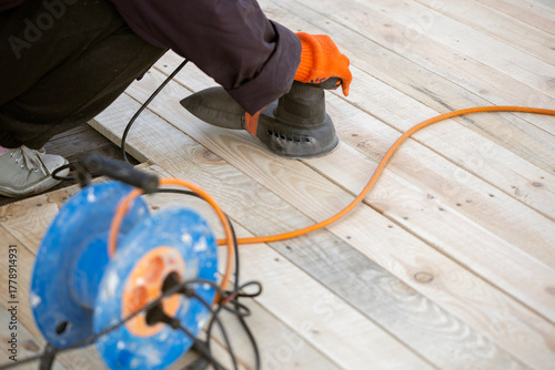 A worker in an orange glove uses an electric orbital sander to smooth a new wooden deck. An orange extension cord and a blue cable reel are on the planks.