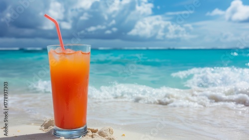 Orange cocktail glass with straw on warm sandy beach beside turquoise water