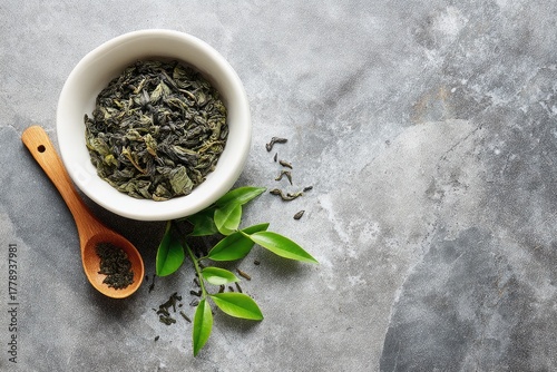 Dried tea leaves in a white bowl with wooden spoon and fresh tea leaves on a gray surface.